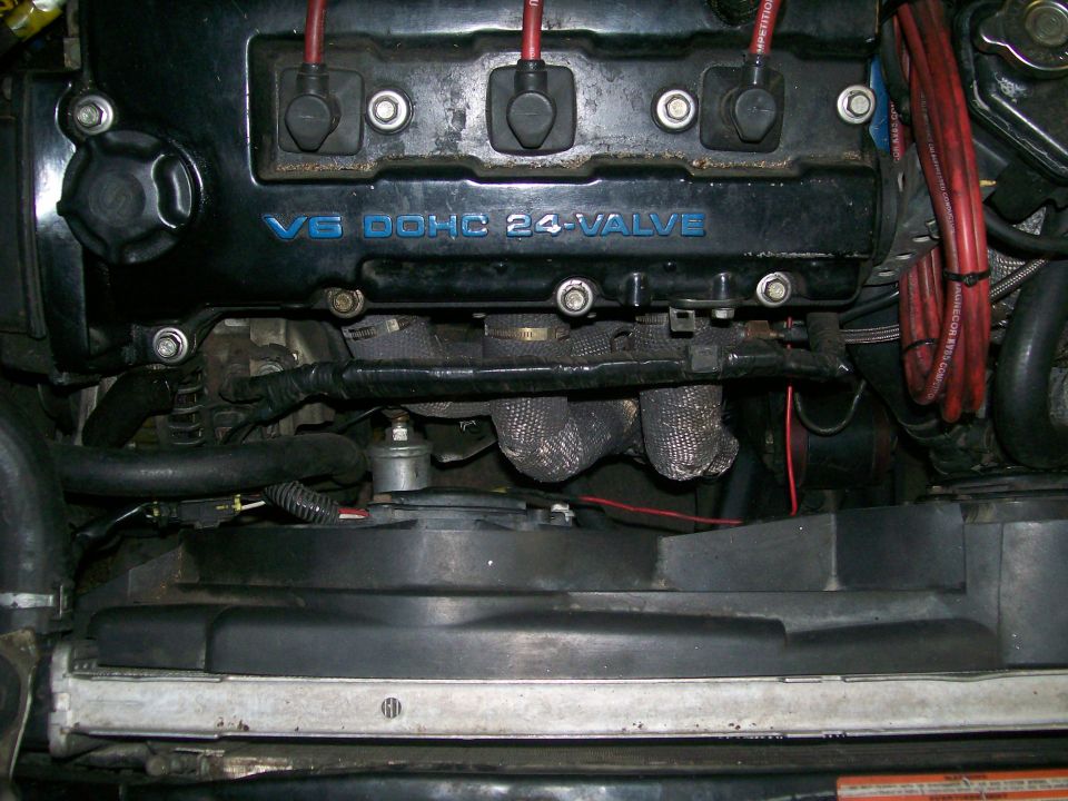 Engine bay with V6 DOHC 24-VALVE text on black cover, exhaust manifold, and fan