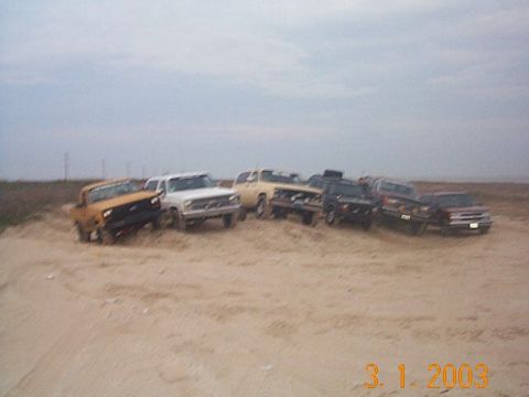 1986 Ford Bronco - A group of trucks and SUVs are parked together on a sandy terrain.