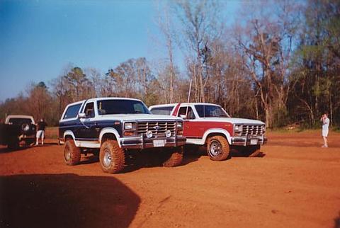1986 Ford Bronco - Two Ford Broncos are parked on a dirt lot with trees in the background. One is blue and white, the other red and white.