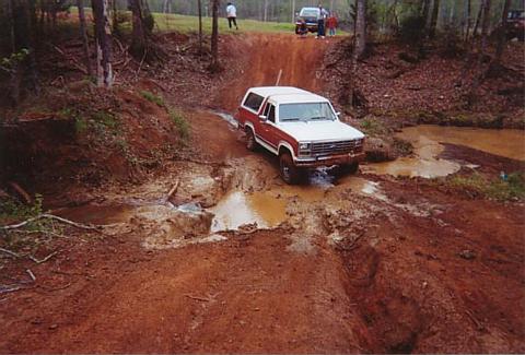 1986 Ford Bronco - A red and white Ford Bronco drives through a muddy obstacle course with other vehicles visible in the background.