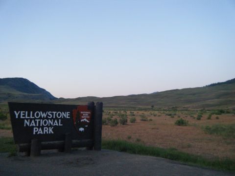 1981 Ford E350 - A sign for Yellowstone National Park stands against a backdrop of rolling hills and a vast, open landscape under a clear sky.