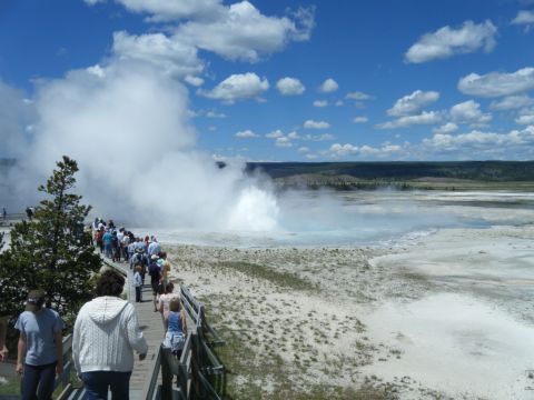 1981 Ford E350 - Tourists gather on a wooden walkway to witness the powerful eruption of a geyser under a blue, cloudy sky.