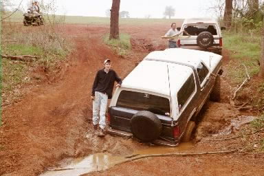 1986 Ford Bronco - Two Ford Broncos navigate a muddy off-road trail with people present.