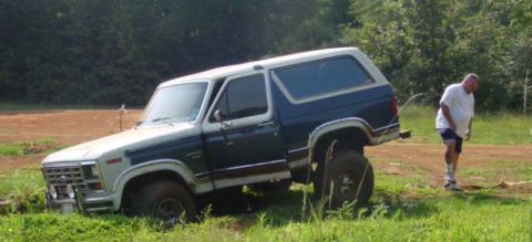 1986 Ford Bronco - A blue and white 1986 Ford Bronco is stuck in a grassy ditch with a man standing nearby.