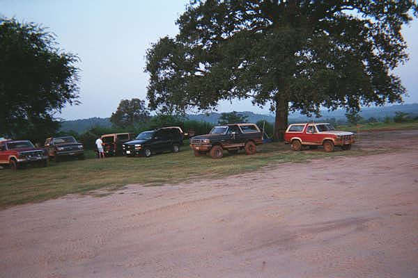 Row of lifted 4x4 trucks and Broncos parked under a tree on a dirt overlook at dusk