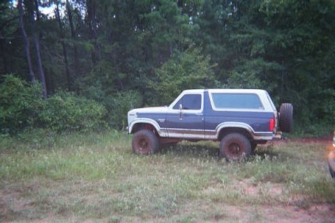 1986 Ford Bronco - A blue and white Ford Bronco is parked in a grassy field with trees behind it.