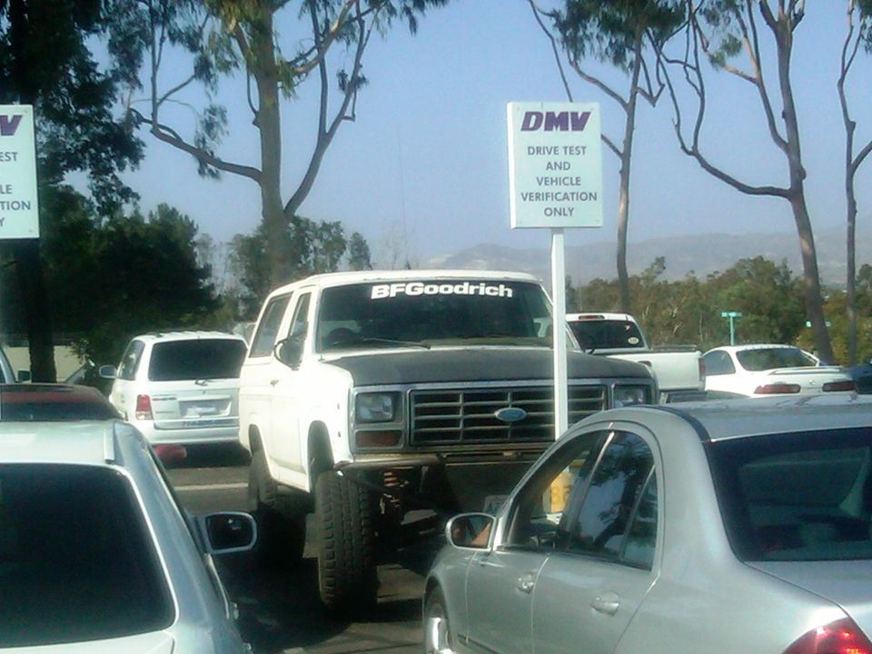 Lifted white Ford Bronco with BFGoodrich sticker on windshield, parked at DMV. Other vehicles visible