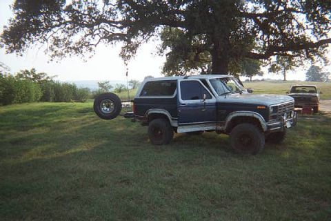 1986 Ford Bronco - A blue and white 1986 Ford Bronco is parked in a grassy area with a spare tire mounted on the rear, and another vehicle is visible in the b…