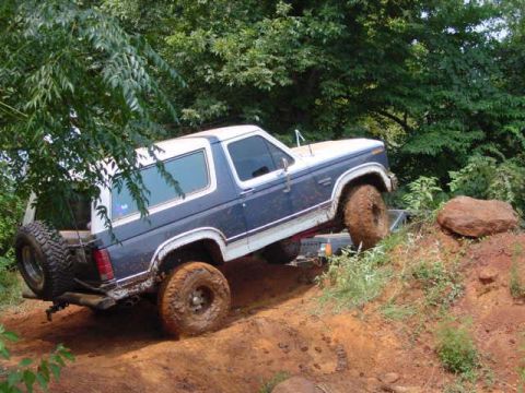 1986 Ford Bronco - A blue and white Ford Bronco navigates a muddy, off-road trail with trees in the background.
