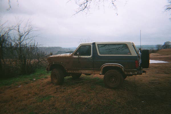 Muddy dark blue Ford Bronco with white hardtop parked on a grassy hill under gray sky