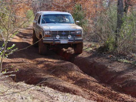1986 Ford Bronco - A silver Ford Bronco drives through a rutted dirt path in a wooded area.