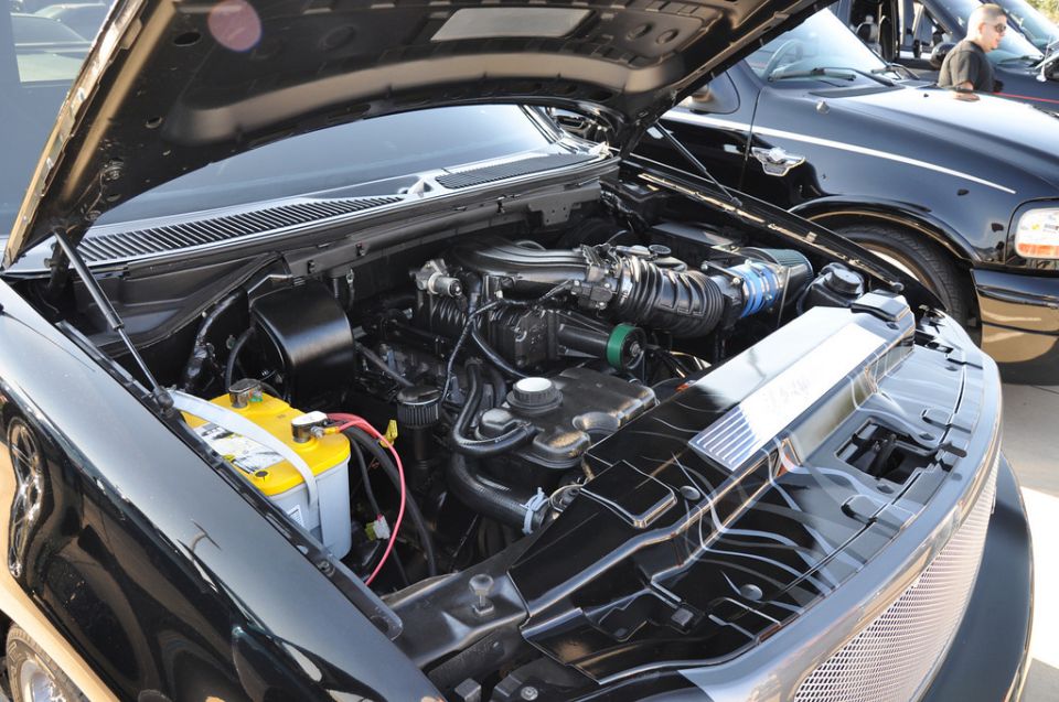 Engine bay of a black truck with hood open, showing engine, battery, and part of another black truck in background