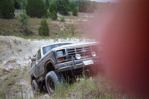 1986 Ford Bronco - A Ford Bronco is shown off-road on a dirt path with grassy terrain and trees in the background.