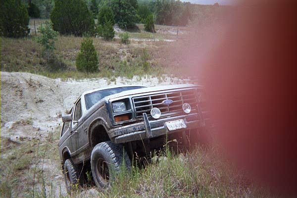 Muddy dark Ford Bronco with brush guard at the edge of a sandy wash, red light leak