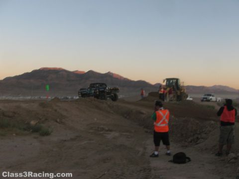 1990 Ford Bronco - off-road race truck launches over a dirt course at dusk while spectators in orange safety vests and a loader look on.