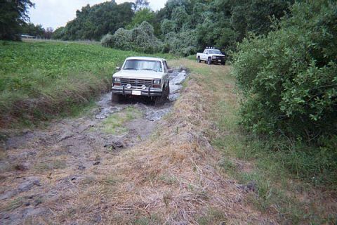 1986 Ford Bronco - Two trucks navigate a muddy trail surrounded by greenery and trees.