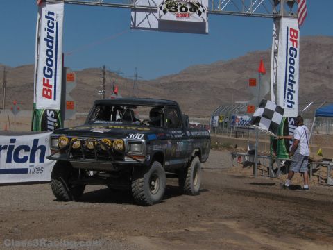 1990 Ford Bronco - black number 300 off-road Ford truck stages at a desert race start line beneath a SCORE banner with a waving checkered flag.