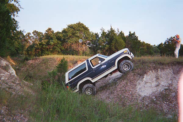 Dark blue and white Ford Bronco climbing a steep dirt embankment with a person standing on the ridge above