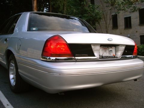 2000 Ford Crown Victoria - The rear of a silver sedan shows its taillights and a black trim panel with 'Crown Victoria' badging.