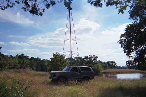 1986 Ford Bronco - A Ford Bronco sits in a grassy field with a windmill and pond in the background under a cloudy sky.