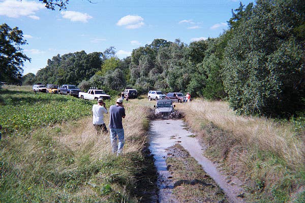 Group of trucks and people gathered at a grassy field trailhead under blue sky