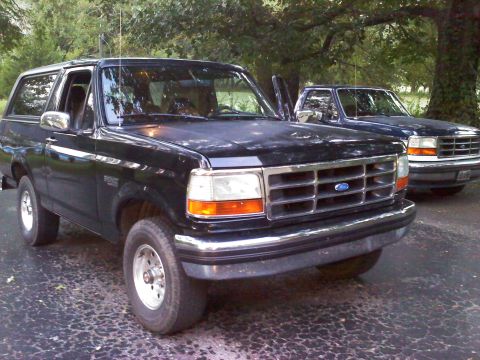 1995 Ford Bronco - Two Ford Broncos are parked on a driveway, one black and one dark blue, with trees in the background.