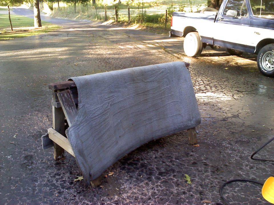 Wet gray carpet drying over a wooden sawhorse on wet pavement, blue Ford truck in background