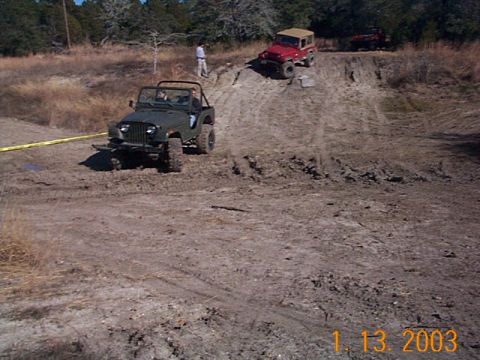 1986 Ford Bronco - Several off-road vehicles navigate a dirt track with steep inclines and muddy patches.
