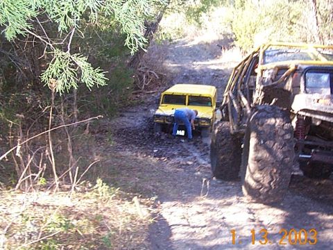 1986 Ford Bronco - A yellow off-road vehicle is stuck in the mud on a trail, with another large off-road vehicle to its right.