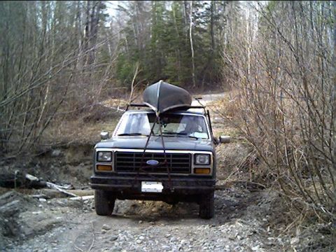 1981 Ford Bronco - A Ford Bronco with a canoe on its roof rack navigates a wooded dirt road.