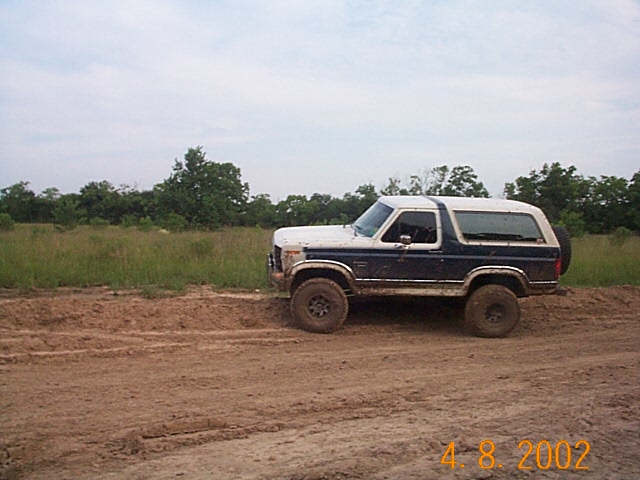 Blue and white Ford Bronco parked on a dirt road with trees and grass in the background