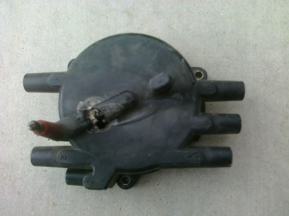 Black plastic distributor cap with four spark plug wire terminals and one ignition coil wire terminal, resting on a concrete surface