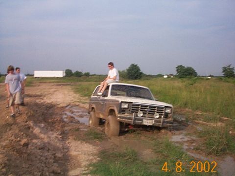1986 Ford Bronco - A Ford Bronco navigates a muddy terrain with a passenger perched atop its roof.