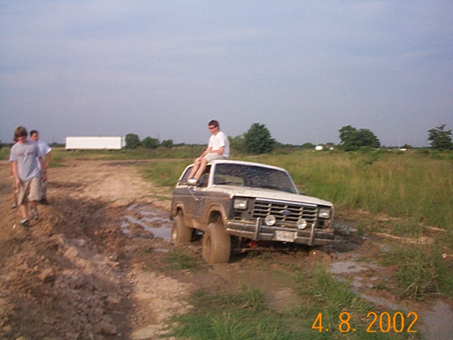 Muddy topless 1980s Ford Bronco in a rutted mud pit with a rider on the roll bar, dated 2002