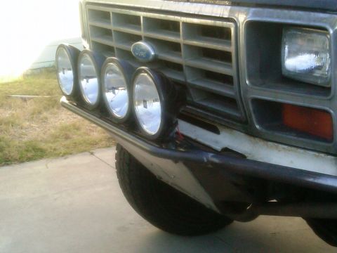 1986 Ford Bronco - Close-up of the front bumper and grille of a 1986 Ford Bronco, four auxiliary lights mounted on a custom bar.