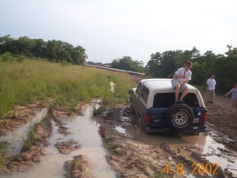 1986 Ford Bronco - A Ford Bronco is stuck in a muddy off-road track, with a person sitting on the spare tire on the back.