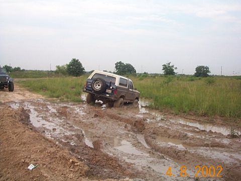 1986 Ford Bronco - A blue and white Ford Bronco is stuck in a muddy off-road trail, with a blue Jeep visible in the background.