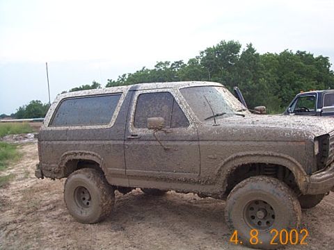 1986 Ford Bronco - A heavily mud-splattered SUV is parked off-road, with another vehicle partially visible behind it.
