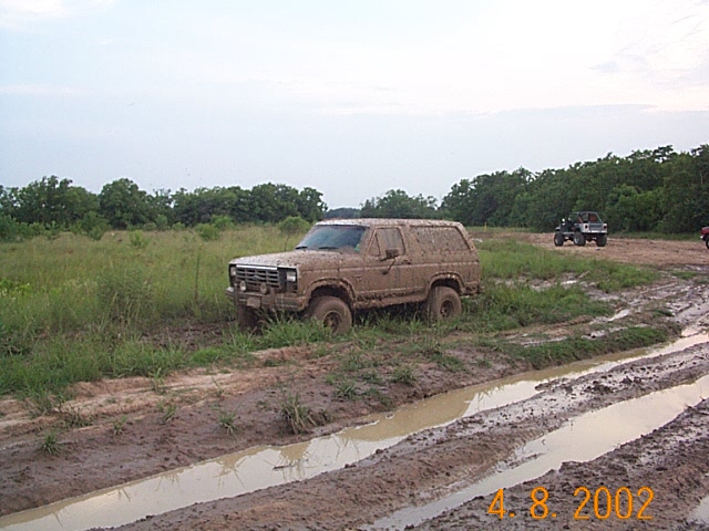 1980s Ford Bronco caked in dried mud beside water-filled ruts in a field, dated 2002