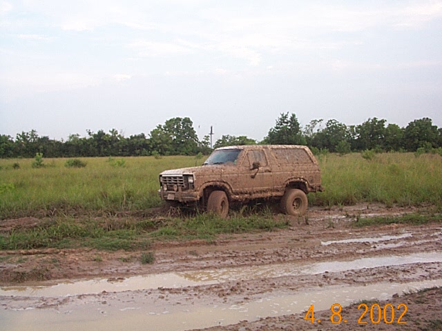 Ford Bronco completely covered in dried mud parked in a grassy field beside water-filled ruts