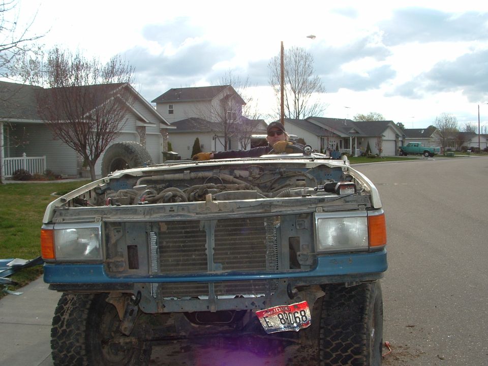 Front of blue Ford with stripped front end, exposed radiator and wiring, Idaho plate, person leaning over it