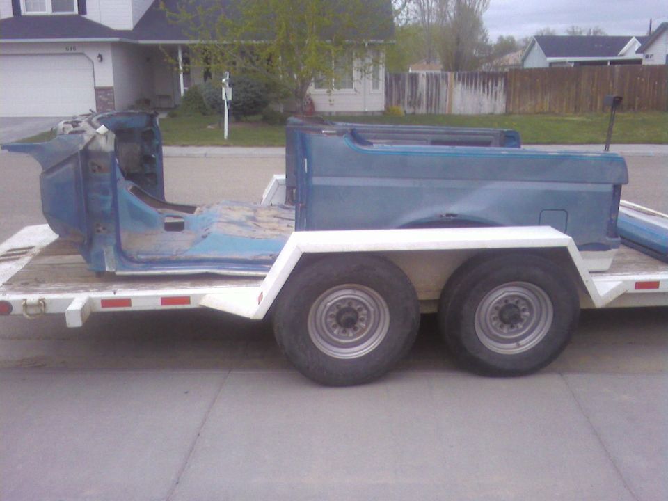 Blue Bronco body shell and rear tub sections on a white tandem-axle trailer parked on a street