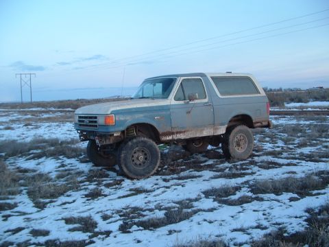 1989 Ford Bronco - A lifted Ford Bronco sits in a field with patchy snow under a pale blue sky.