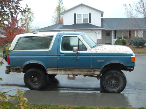 1989 Ford Bronco - A muddy blue and white Ford Bronco is parked on a wet road in front of a house.
