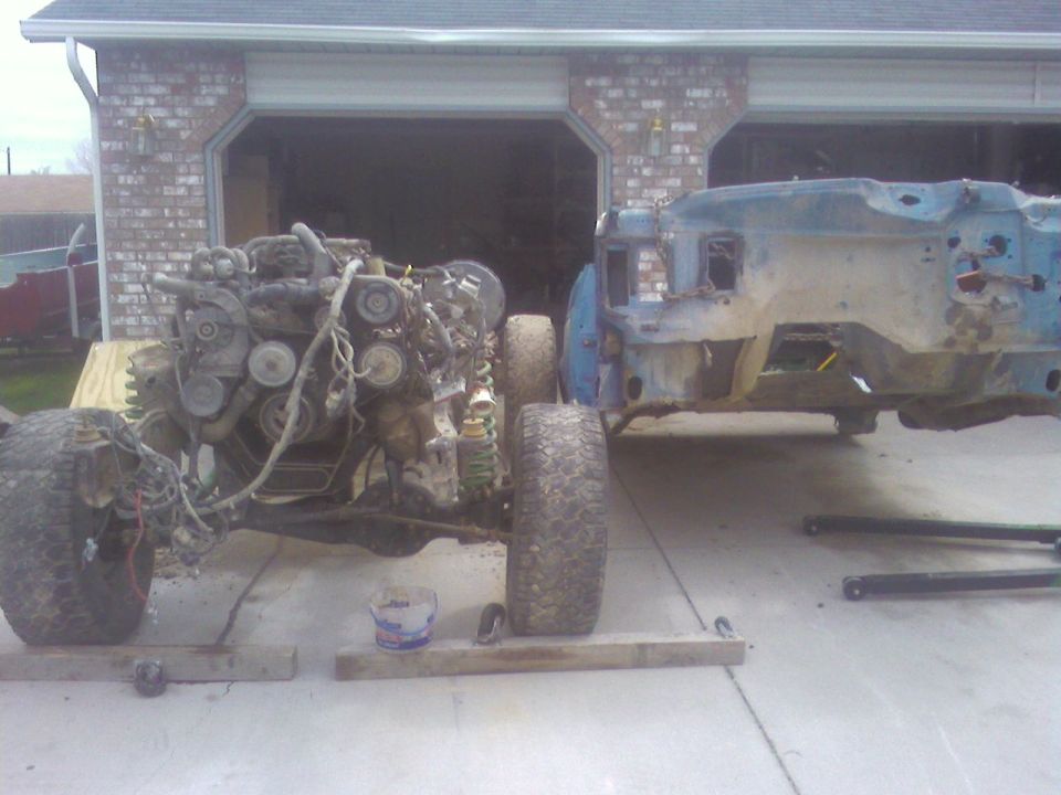 Truck frame with engine on rolling cart beside blue body shell in driveway