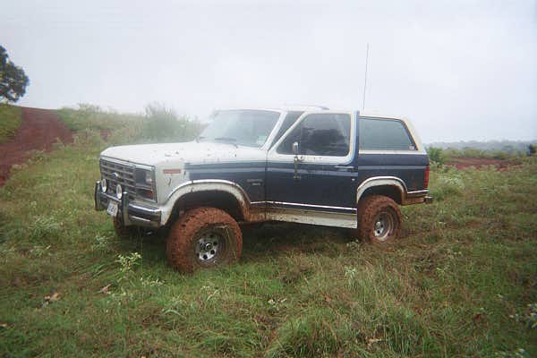 Blue and white Ford Bronco with muddy tires parked in foggy grass field