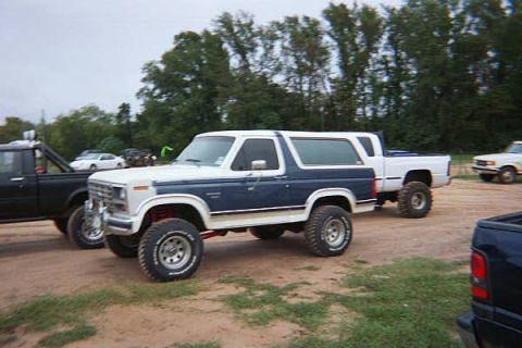 1986 Ford Bronco - A white and blue Ford Bronco is parked on a dirt lot next to other vehicles.