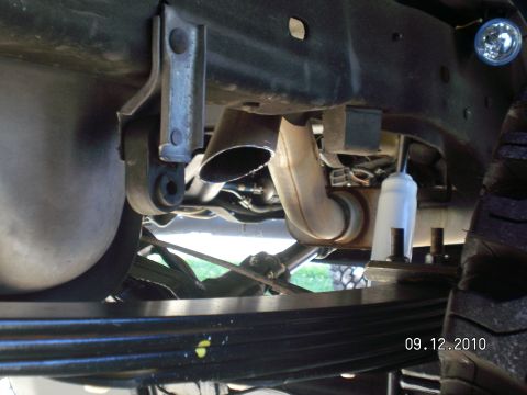 1996 Ford Bronco - Underbody view along a truck frame rail showing an angle-cut exhaust tailpipe exiting ahead of the rear tire, a hanger bracket, and the fue…