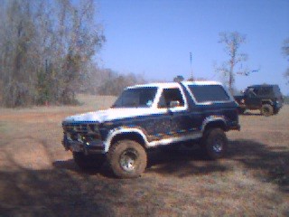 1986 Ford Bronco - A white and blue Ford Bronco from the 1980s is seen in an off-road setting with another vehicle in the background.