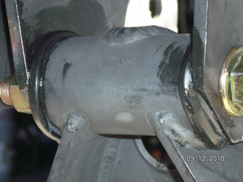 1996 Ford Bronco - Tight underbody close-up of a welded steel radius arm end pivoting in a black bushing between the ears of a drop bracket, secured by a yell…